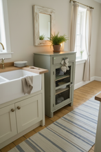 Load image into Gallery viewer, A rustic sage green cabinet with wire-front shelves stands beside a white farmhouse sink in a bright kitchen. A distressed wood mirror hangs on the wall above, with a small potted plant on top and a striped rug on the floor.
