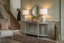 Load image into Gallery viewer, A rustic light gray console table in a styled room, decorated with two matching white table lamps, a round wooden mirror, a small potted plant, and books. A woven armchair is partially visible on the left.
